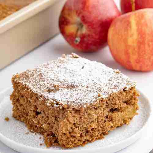 a single slice of apple oatmeal cake dusted with powdered sugar with the cake pan and apples in the background