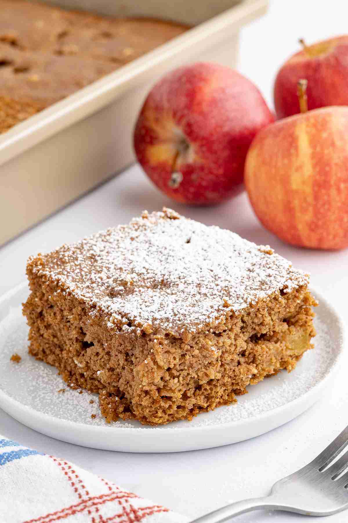 a single slice of apple oatmeal cake dusted with powdered sugar with the cake pan and apples in the background