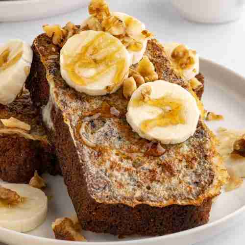 two slices of banana bread french toast on white plate with banana nuts and syrup