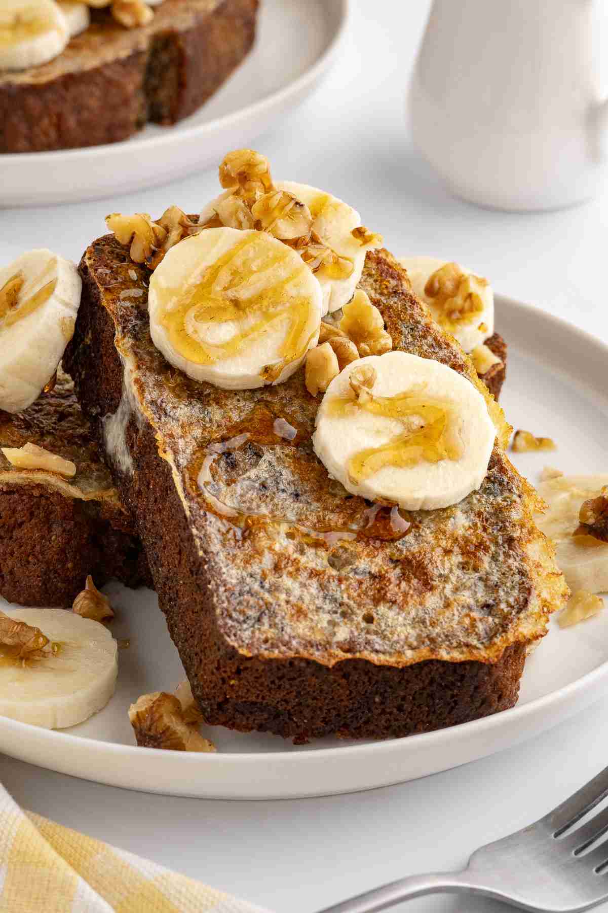 two slices of banana bread french toast on white plate with banana nuts and syrup