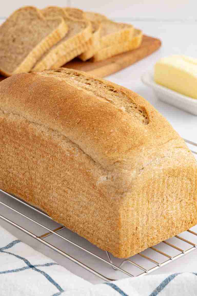 a loaf of fresh milled flour bread on cooling rack with sliced bread and butter in the background