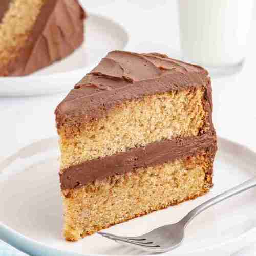 a slice of fresh milled yellow cake with chocolate frosting on white plate with milk and another slice in the background