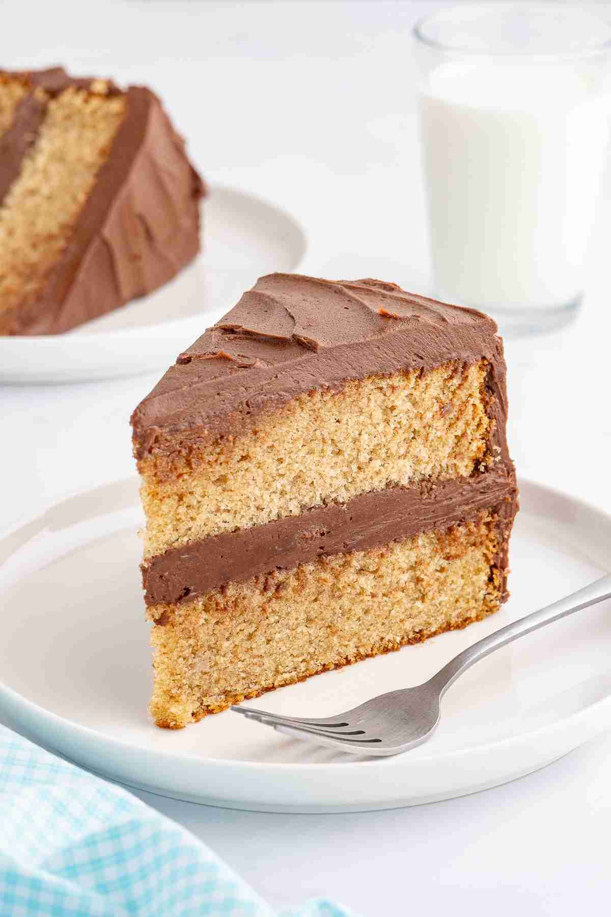 a slice of fresh milled yellow cake with chocolate frosting on white plate with milk and another slice in the background