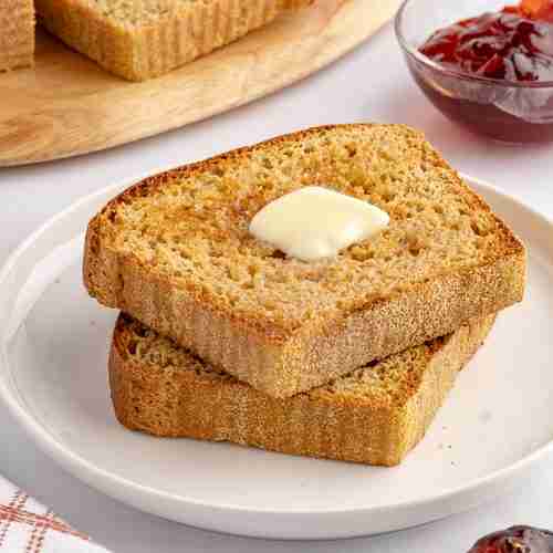 two slices of fresh milled English muffin toasting bread with butter on serving plate with jelly and more bread in the background