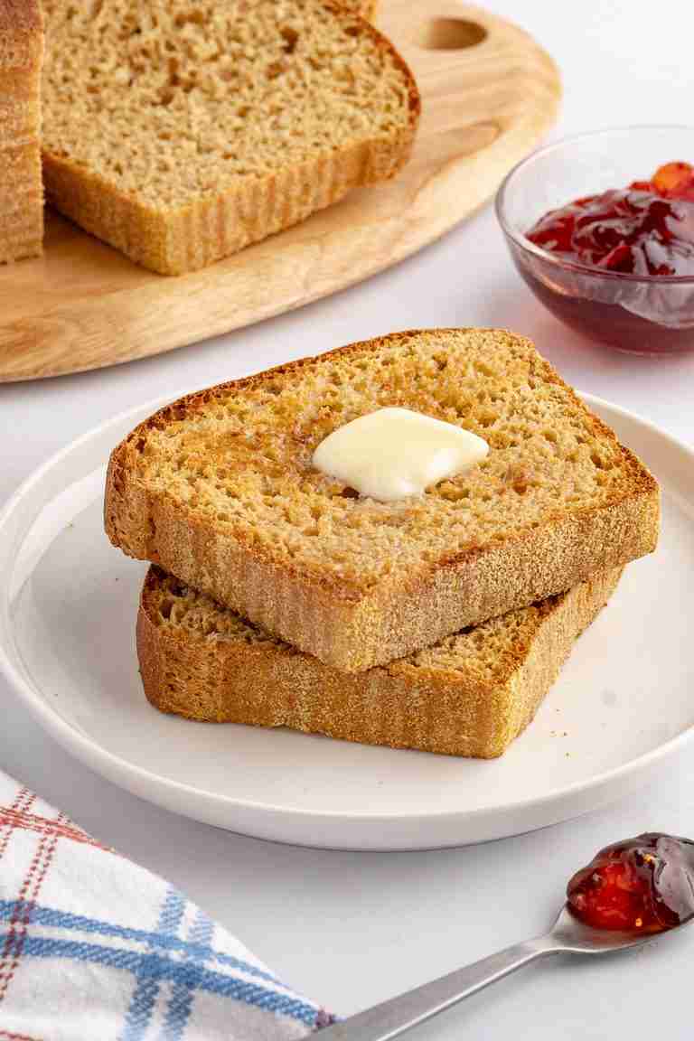 two slices of fresh milled English muffin toasting bread with butter on serving plate with jelly and more bread in the background