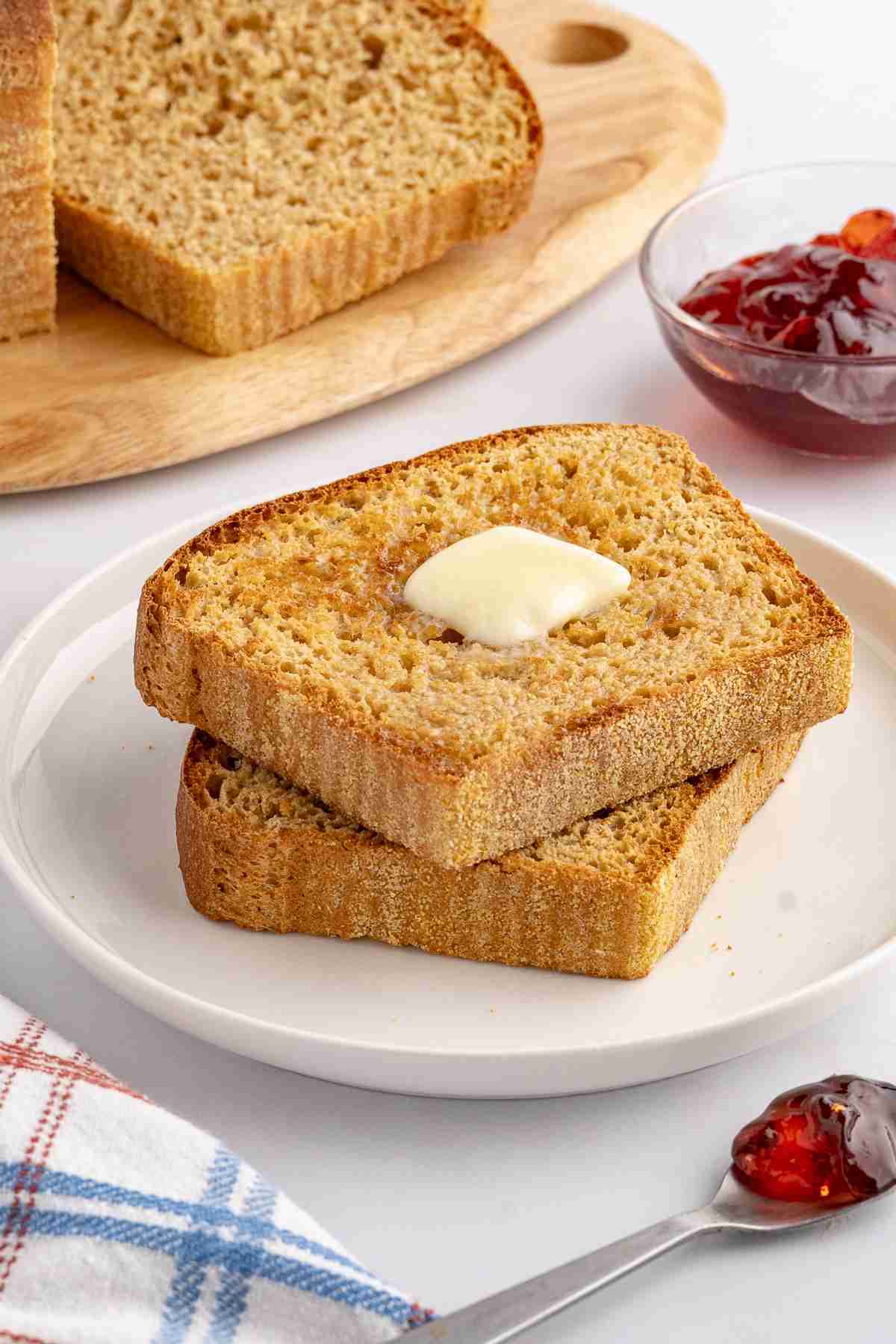 two slices of fresh milled English muffin toasting bread with butter on serving plate with jelly and more bread in the background
